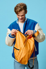 a student in a blue bomber jacket looks into his orange backpack while standing against a blue...