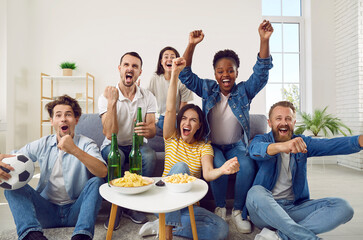 Happy diverse male and female friends enjoying weekend and watching soccer on TV. Group of young multiracial people sitting on sofa, watching football, celebrating goal, raising hands up and screaming