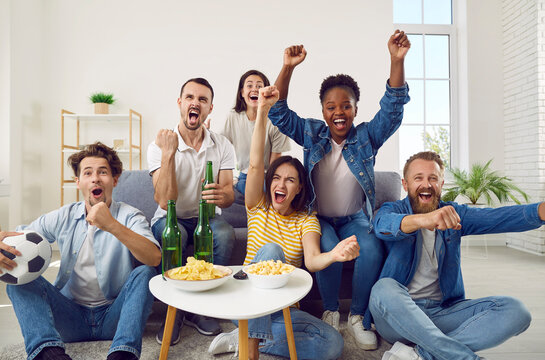 Happy Diverse Male And Female Friends Enjoying Weekend And Watching Soccer On TV. Group Of Young Multiracial People Sitting On Sofa, Watching Football, Celebrating Goal, Raising Hands Up And Screaming