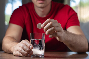A man takes a cure for a hangover at home, throws a second fizzy pill into a glass, focusing on the hand with the pill