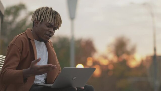 Online meeting. Inspired man. Video communication. Hipster black guy sitting square bench talking on laptop in sunset light.