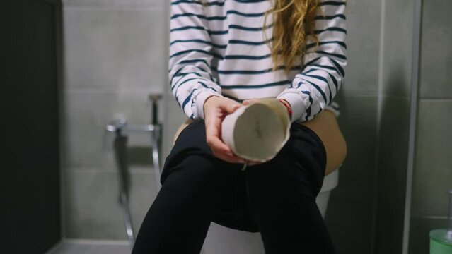 Low section of woman body sitting on toilet and suffers from diarrhea and stomachache. Female can't poop because of hemorrhoids piles. Strained girl holding and shaking toilet paper in hands.