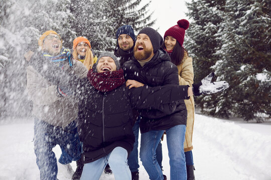 Team of happy young friends having lots of fun in the snow. Group of cheerful adult people in warm hats, coats and jackets walking together in a winter forest, throwing snow in the air and laughing