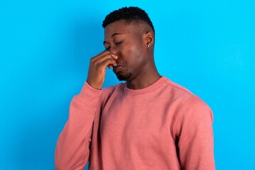 young handsome man wearing pink sweater over blue background smelling something stinky and disgusting, intolerable smell, holding breath with fingers on nose. Bad smell