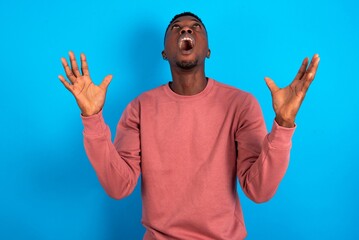 young handsome man wearing pink sweater over blue background crazy and mad shouting and yelling with aggressive expression and arms raised. Frustration concept.