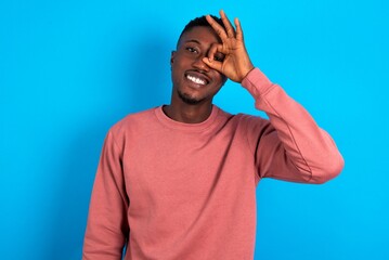 young handsome man wearing pink sweater over blue background doing ok gesture with hand smiling, eye looking through fingers with happy face.