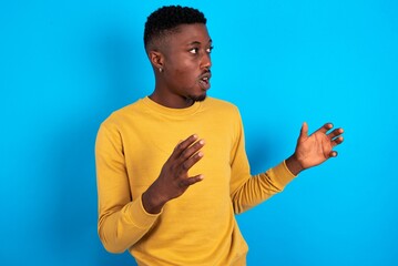 young handsome man wearing yellow sweater over blue background shouts loud, keeps eyes opened and hands tense.