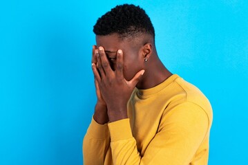 Sad young handsome man wearing yellow sweater over blue background crying covering her face with her hands.