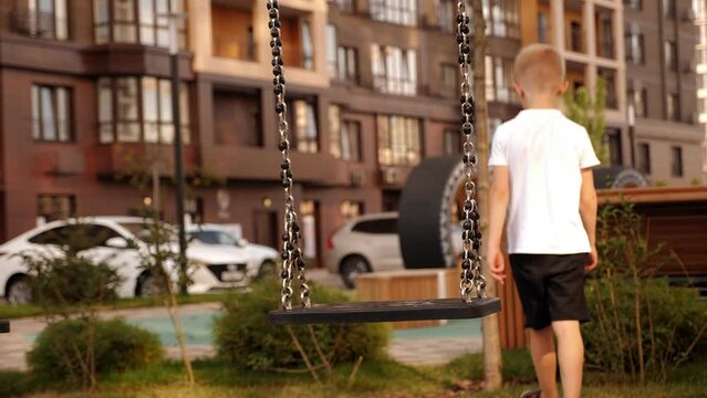 A sad little boy is walking alone in the yard on a modern playground, he rode on a swing and now leaves alone. The child is a loner without friends.