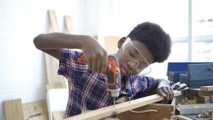 A young african boy wearing shirt and using screwdriver and try to screw the wood piece in the classroom. Smiling kid carpenter happy working with wood and sandpaper, preschool and learning concept.
