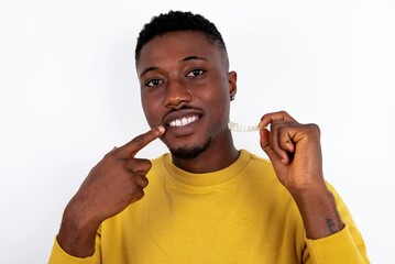 young handsome man wearing yellow sweater over white background holding an invisible aligner and...