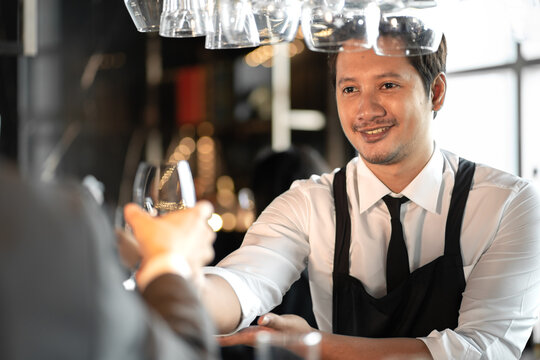 Smiling male bartender working in nightlife serving a glass of alcohol cocktail drink in pub. Asian barkeeper standing at counter bar to service liquid beverage or whiskey for refreshment in nightclub