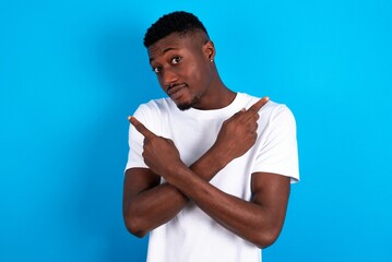 young handsome man wearing white T-shirt over blue background crosses arms and points at different sides hesitates between two items or variants. Needs help with decision