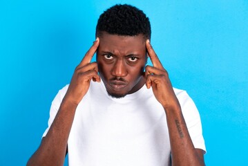 Serious concentrated young handsome man wearing white T-shirt over blue background keeps fingers on temples, tries to ease tension, gather with thoughts and remember important information for exam