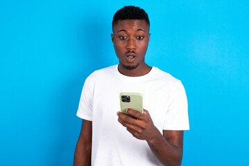 young handsome man wearing white T-shirt over blue background holds mobile phone in hands and rejoices positive news, uses modern cellular