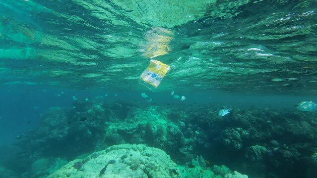 Slow motion, discarded plastic packaging slowly drifting under the surface of water nearby coral reef a tropical fish swimming around.  