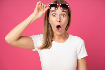 impressed shocked young attractive woman dressed in white t-shirt and holding sunglasses on her head with open mouth omg and big eyes isolated over pink red background - obrazy, fototapety, plakaty