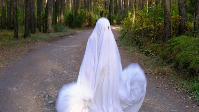 A child in sheets with cutout for eyes like a ghost costume dancing in an autumn forest scares and terrifies. A kind little funny ghost. Halloween Party, slow motion 4k