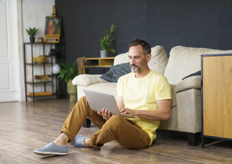 Handsome man using laptop sitting on floor at leaving room lean on sofa learning, working, watching video, movie or webinar.