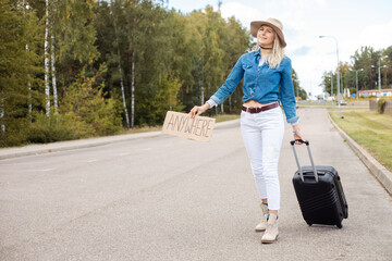 Joyful woman wait passing car on empty road with cardboard poster and suitcase. Lady in hat and...