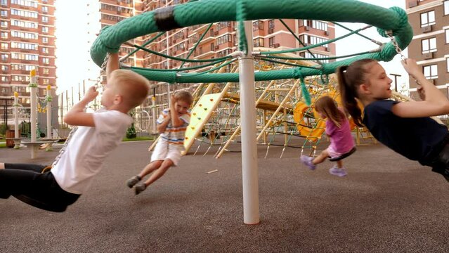 Four children ride a carousel sitting on ropes with seats on the playground in a modern residential complex.