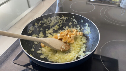 Preparing farofa (Brazilian toasted cassava flour) by toasting in a pan on the electric stove. Bananas, onions, and garlic are fried in a pan while stirred by a wooden spoon.