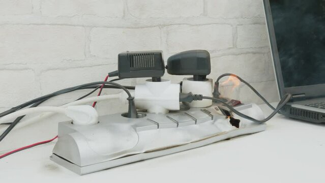 Overloaded power cube on fire on table indoors close up, voltage overload. Lot of electric plugs in sockets of power strip.