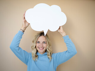 beautiful young woman with curly blond hair stands in front of brown wall and holds white speech...