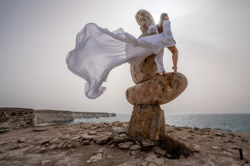 A woman stands on a stone sculpture made of large stones. She is dressed in a white long dress,...