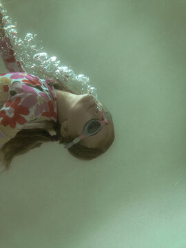 Young girl swimming underwater in pool wearing floral swim suit and goggles blowing bubbles