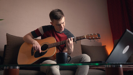 Obraz premium Young man learning to play acoustic guitar online at home.