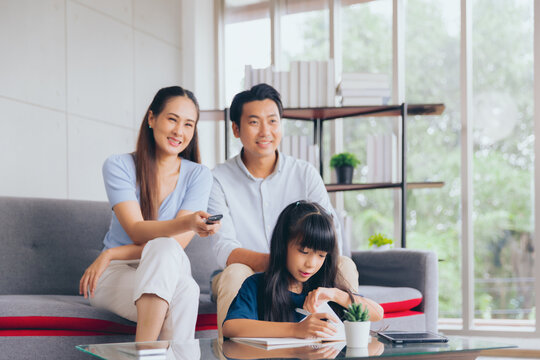 Cheerful asian parent dad and mom watching TV with daughter studying doing homework, Education concept