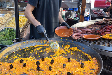Man wearing black gloves dishing up seafood paella from large pan into bowl at food market in Torrevieja, Spain
