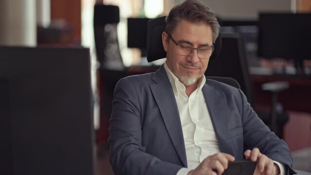 Business portrait - confident businessman sitting at desk working in office, using phone. Happy mid adult man in shirt and jacket, smiling. Bearded, glasses.