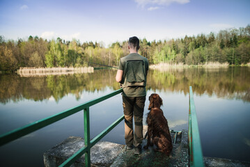 A young man in a vest is fishing on a small pond with a hunting dog. Catching fish in nature at...