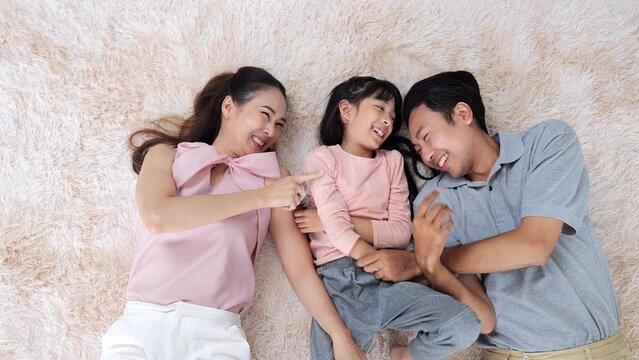 Happy asian family, young father, mother and daughter adorable lay together on the carpet inside living room Tease and tickle each other Laughing happily, looking warm close relationship.