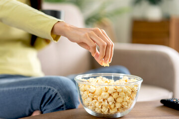 Young asian woman is grabbing popcorn in a bowl and holding coffee cup while sitting on the big comfortable