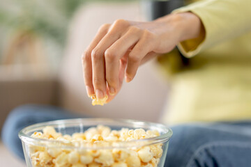 Young asian woman is grabbing popcorn in a bowl and holding coffee cup while sitting on the big comfortable