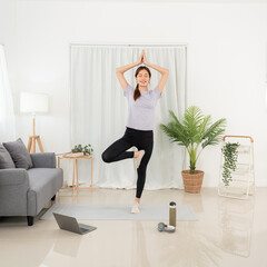 Young woman is watching yoga lesson online on laptop to doing with vrksasana or tree pose while workout