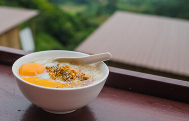 Horizontal picture top view, pork porridge with eggs in a white plate for breakfast, placed on a brown table. There is a blurred background of a view homestay,  in holiday