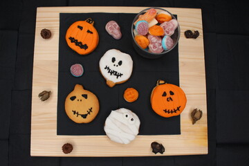 Spooky Halloween cookies with the shape of ghosts and pumpkins. Colors white, brown and orange. They're on a black tissue paper and light wood table. There's a glass full of fun candies and swets.