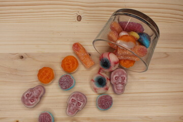 Halloween treats and sweets spill out of a glass tumbled onto a light wooden board. Snack for a family and children's spooky party. They have blue, red and orange colors. Shapes of eyes, skulls, brain
