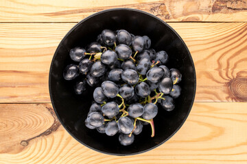 One sprig of sweet black grapes in a black ceramic plate on a wooden table, macro, top view.