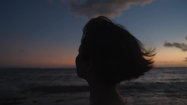 Silhouette side portrait of girl dreaming at seaside in evening light. Woman relaxing meditating alone on ocean beach at colorful sunset sky background. Stress overcome. Film grain texture. Motion