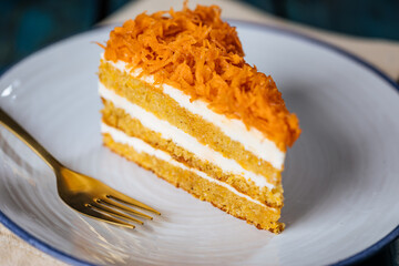 Carrot cake with cream cheese in layers and carrots on top on a  blue and white background with carrots in blurry background