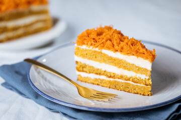 Carrot cake with cream cheese in layers and carrots on top on a  blue and white background with carrots in blurry background