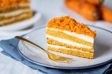 Carrot cake with cream cheese in layers and carrots on top on a  blue and white background with carrots in blurry background