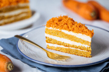 Carrot cake with cream cheese in layers and carrots on top on a  blue and white background with carrots in blurry background