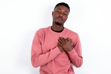 young handsome man wearing pink sweater over white  closes eyes and keeps hands on chest near...