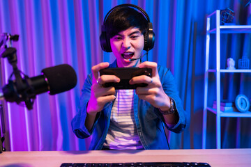 Playing video games on smartphone. Young asian handsome man sitting on chair holding cellphone. Exited streamer wearing headset in online mobile game in neon room.Esport streaming game online.